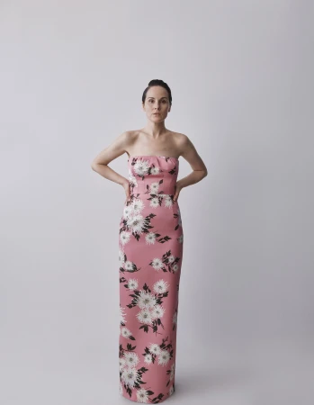 Adult female posing in a long, strapless pink gown with white floral pattern against a plain background in a studio setting.