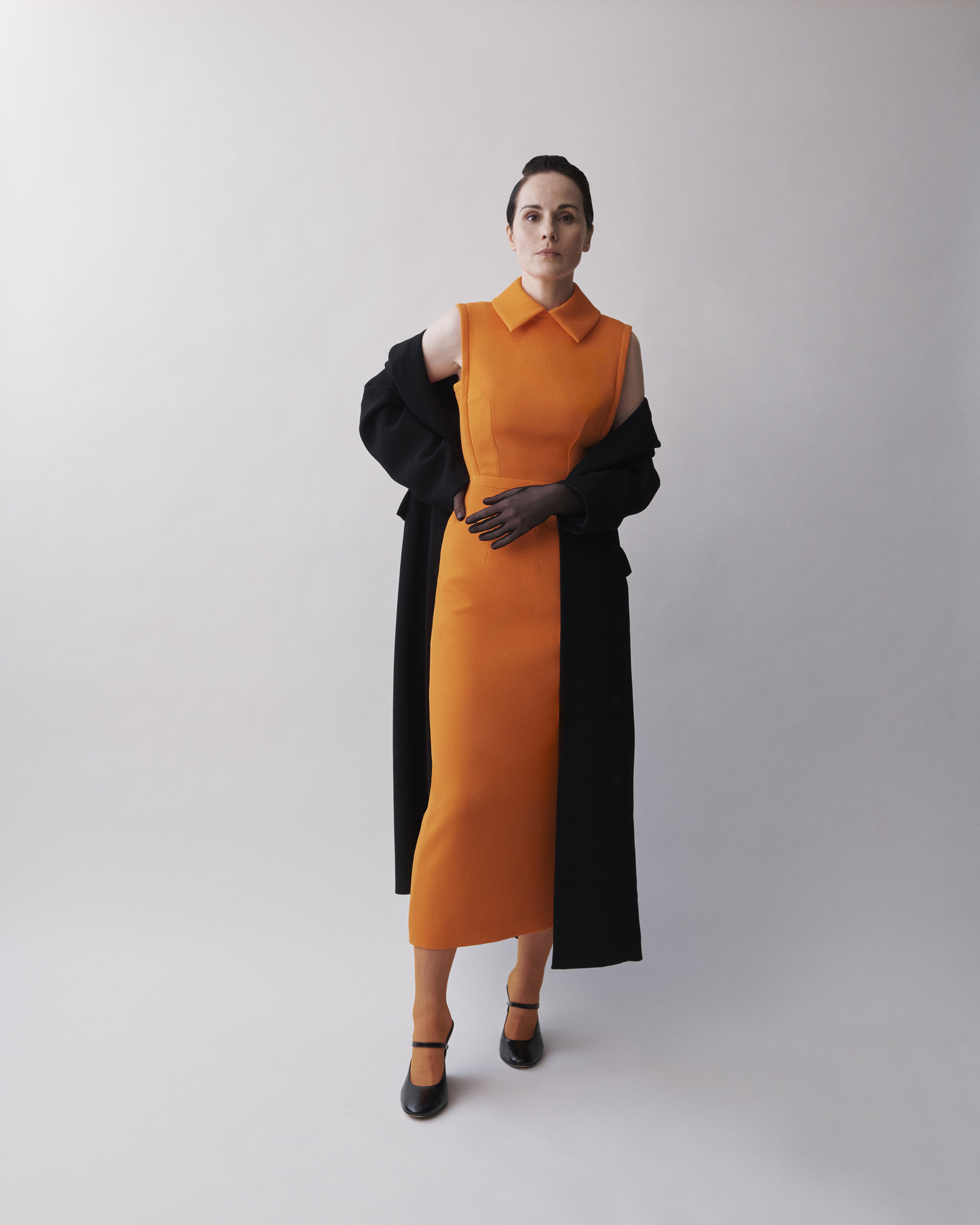 Elegant adult woman in an orange dress and black coat poses against a bright backdrop in a full shot, fashion photography.