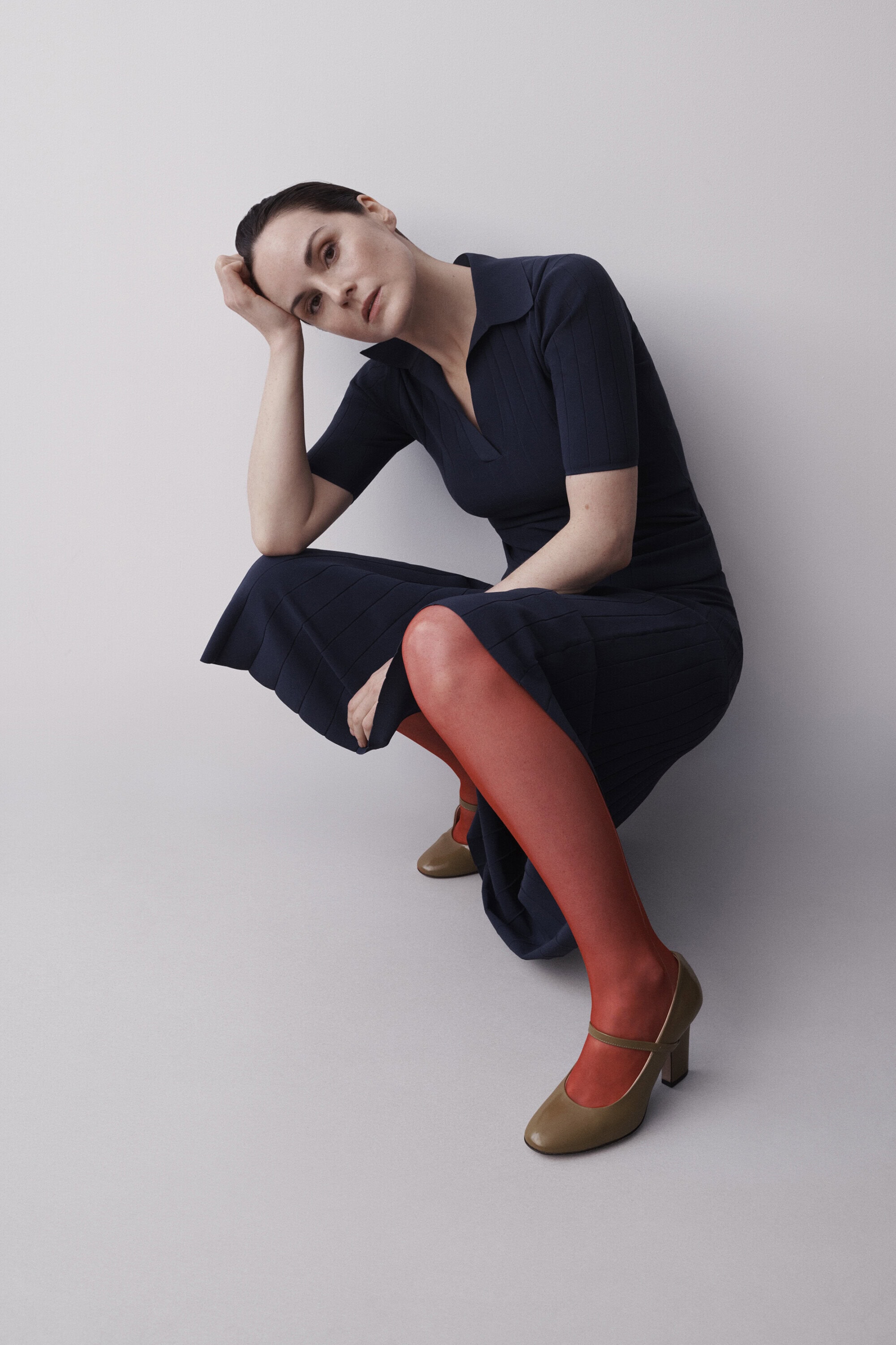 A woman in a navy dress and red tights poses in a studio environment, leaning on her hand with a slightly melancholic expression.