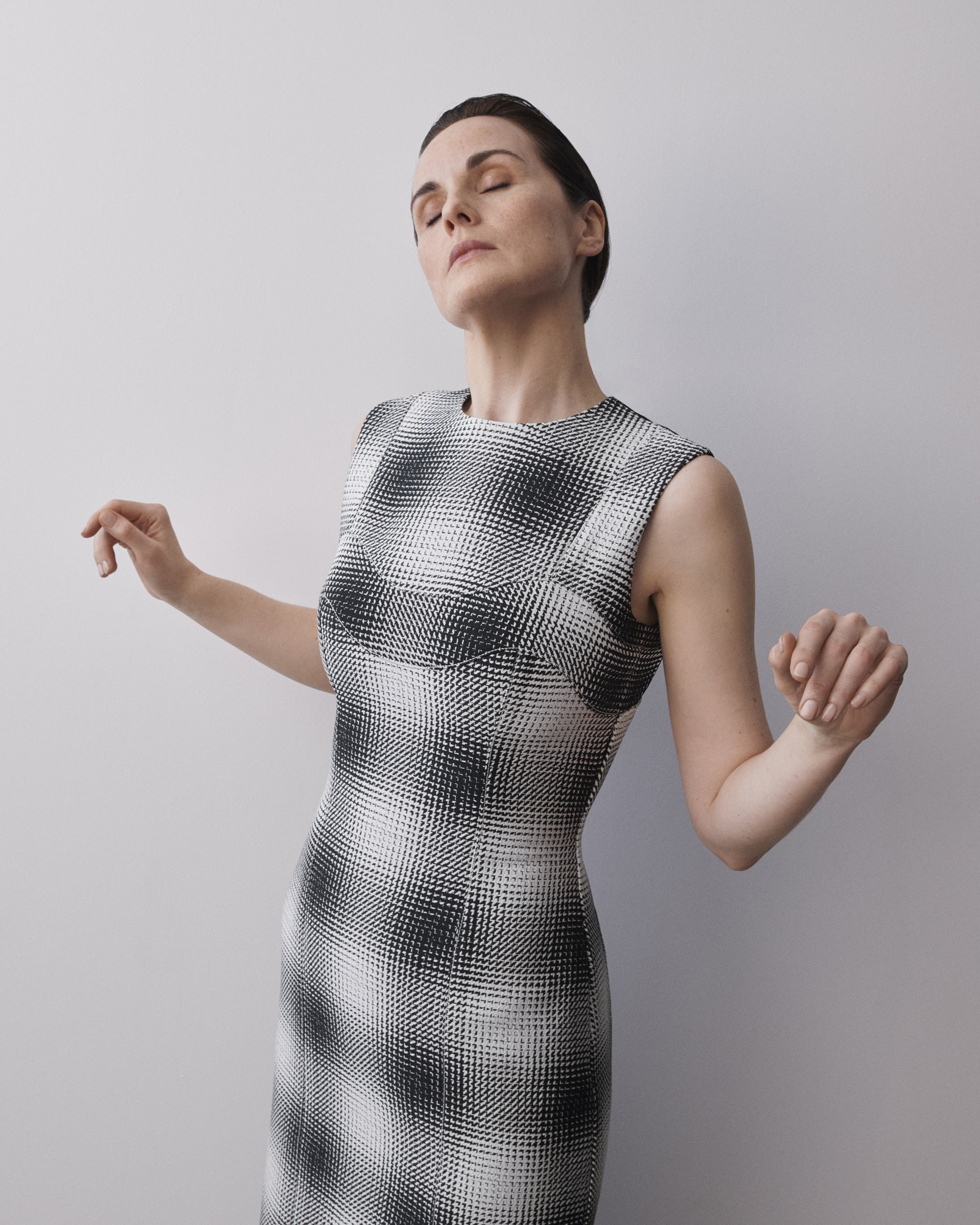 Elegant woman in a black and white patterned dress with closed eyes and arms outstretched against a plain background in a studio setting.