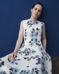 Michelle Dockery wearing a sleeveless white dress with blue floral print against a solid dark blue wall.