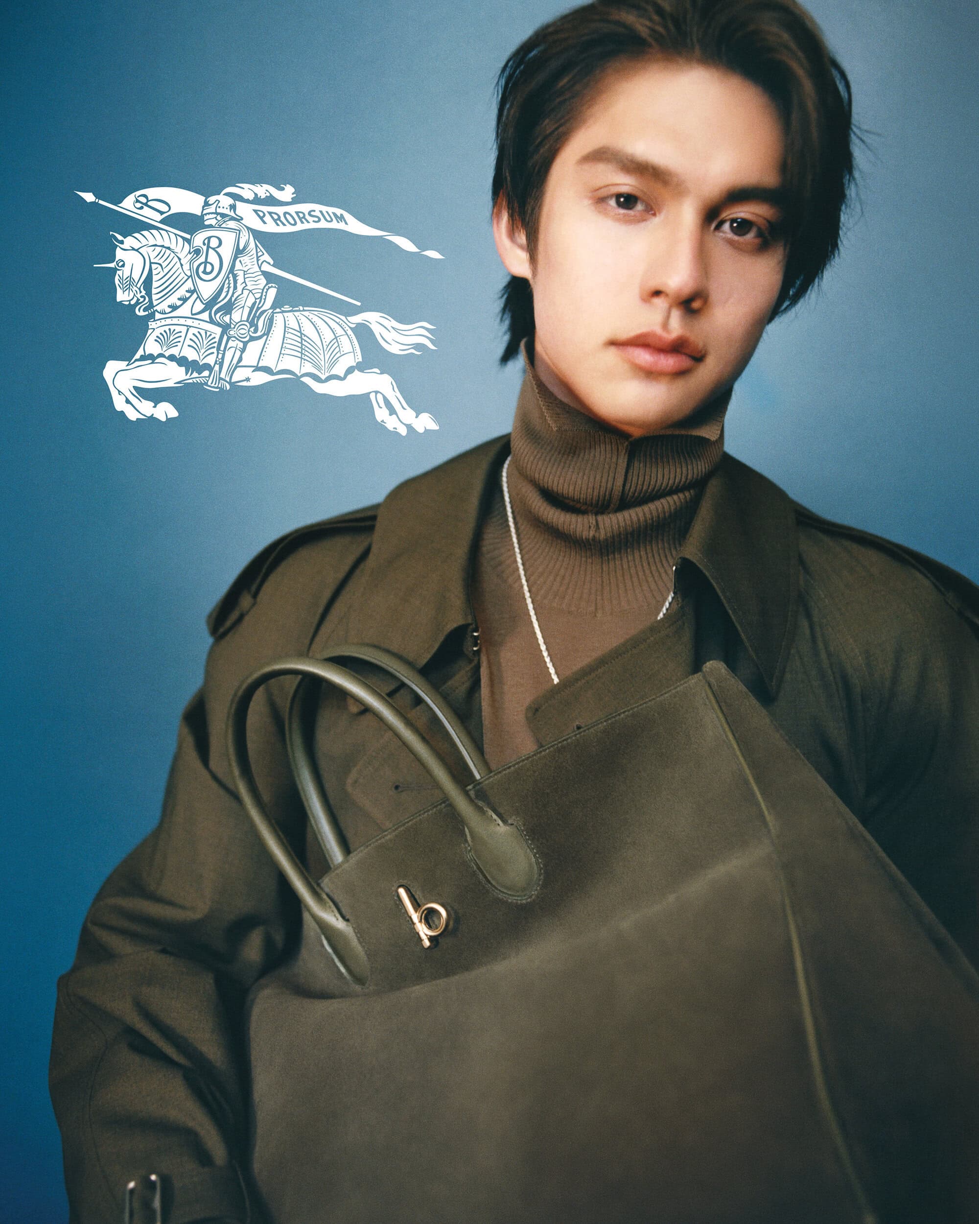 Male model poses indoors wearing a trench coat and carrying a large tote bag, set against a blue background with a logo.