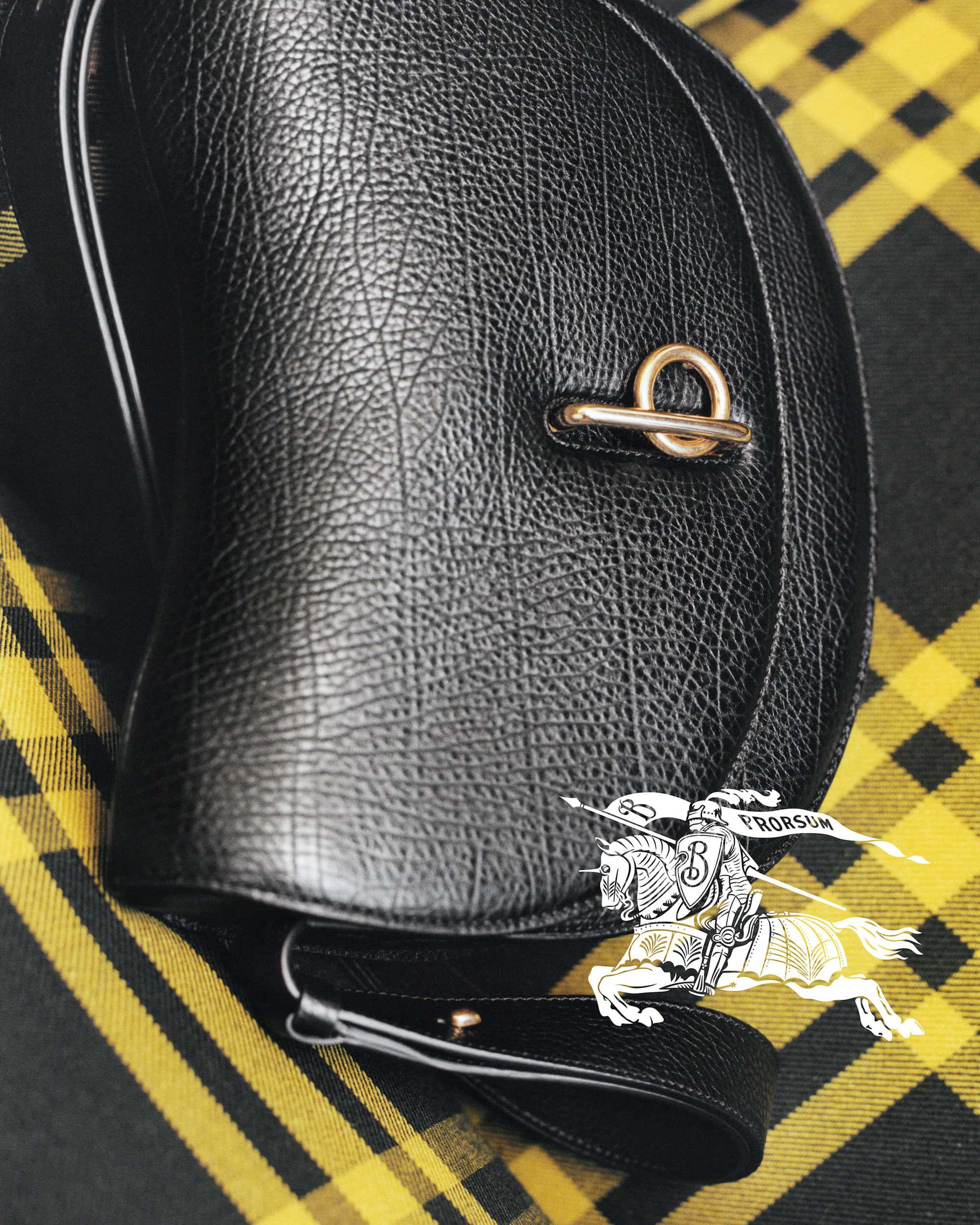 Close-up shot of a black leather handbag with gold hardware on a yellow plaid textile backdrop, featuring the Burberry logo.