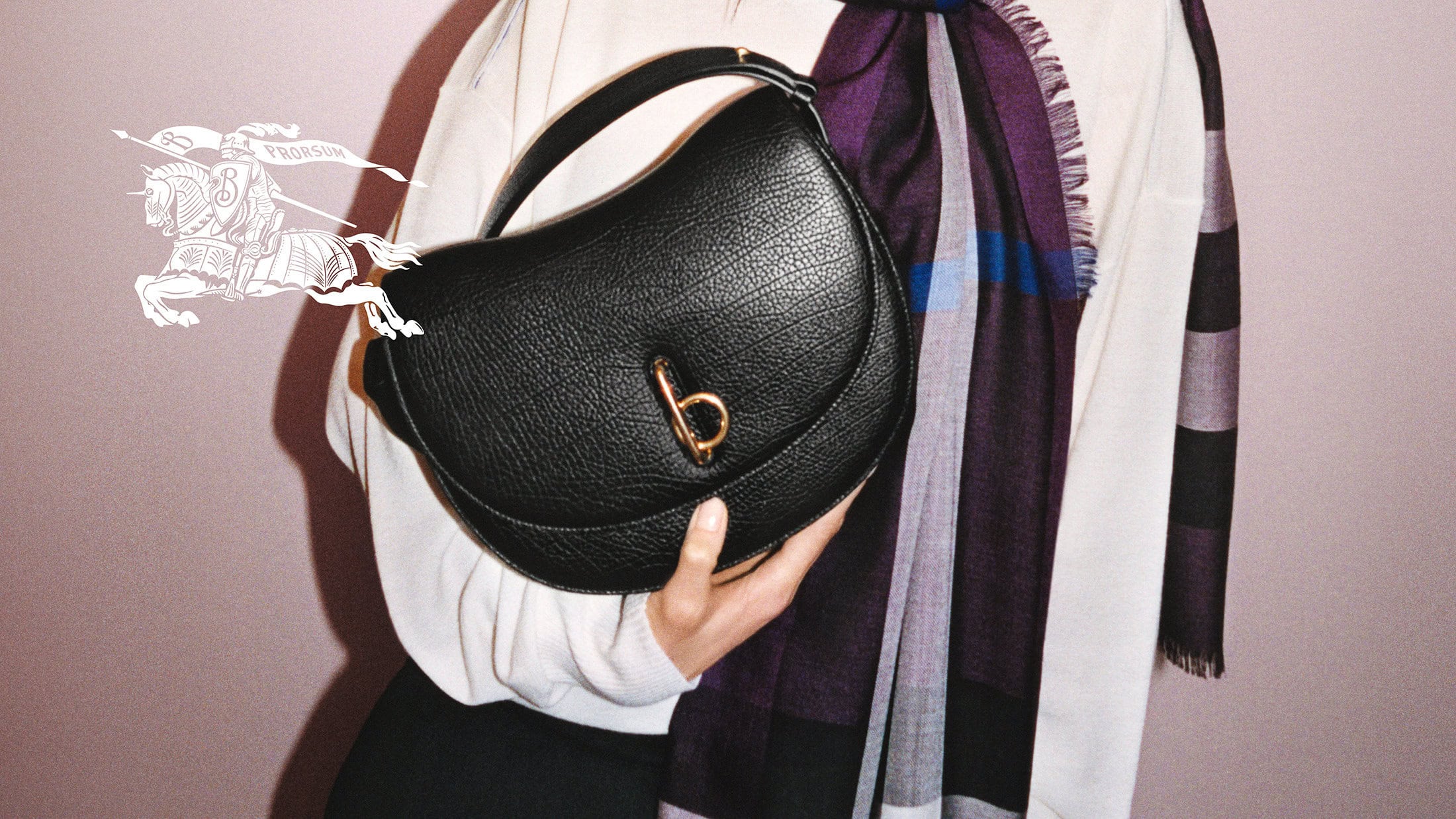 Close-up of a model holding a black leather purse with a gold clasp; purple plaid scarf drapes in the background against a light pink wall.
