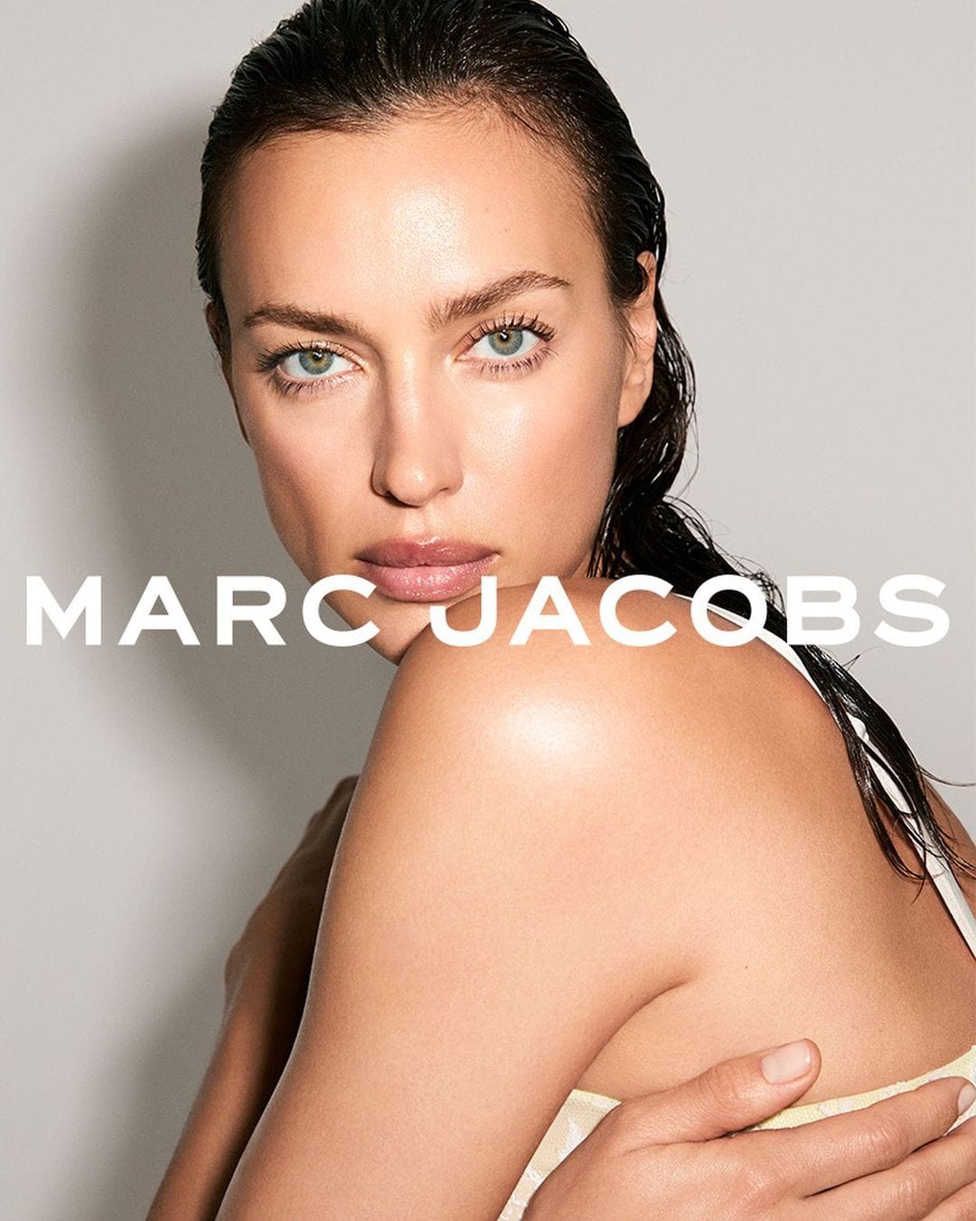 Close-up headshot of Irina Shayk with wet hair and natural makeup, looking at the camera against a neutral background with Marc Jacobs logo.