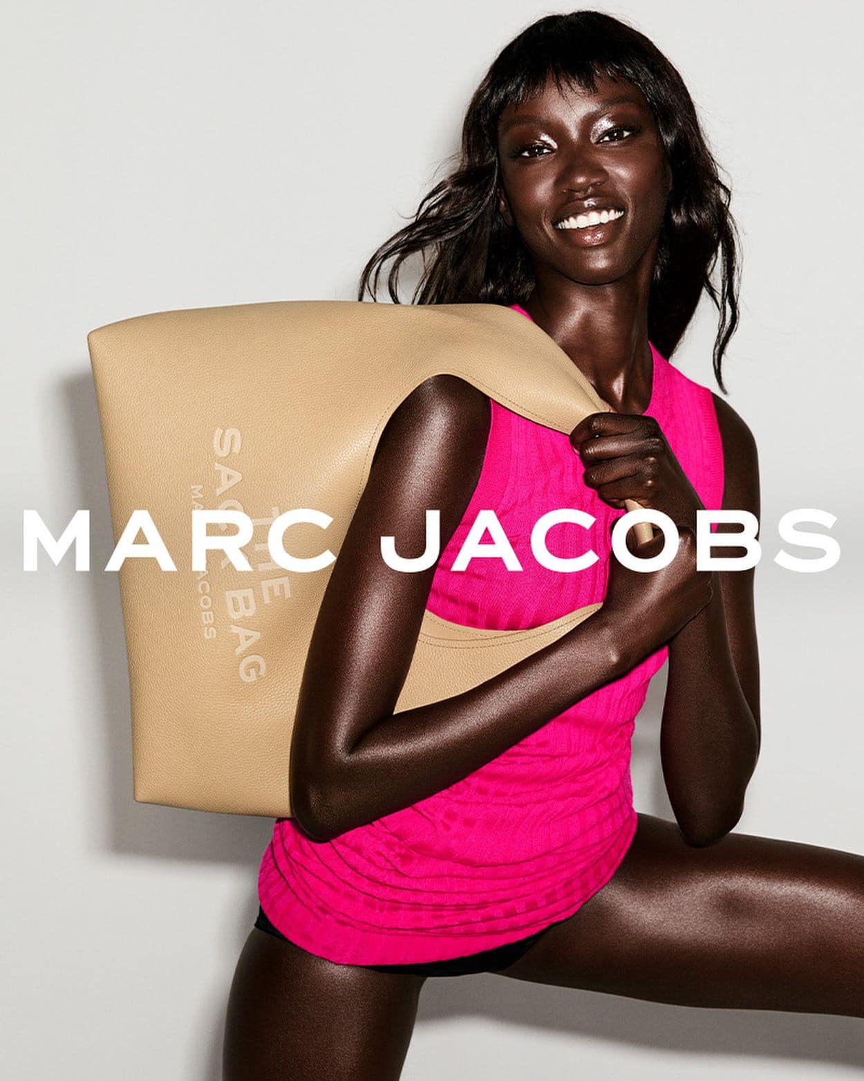 African American woman poses with a Marc Jacobs tote bag. She is wearing a pink top and black bottoms in a studio setting.