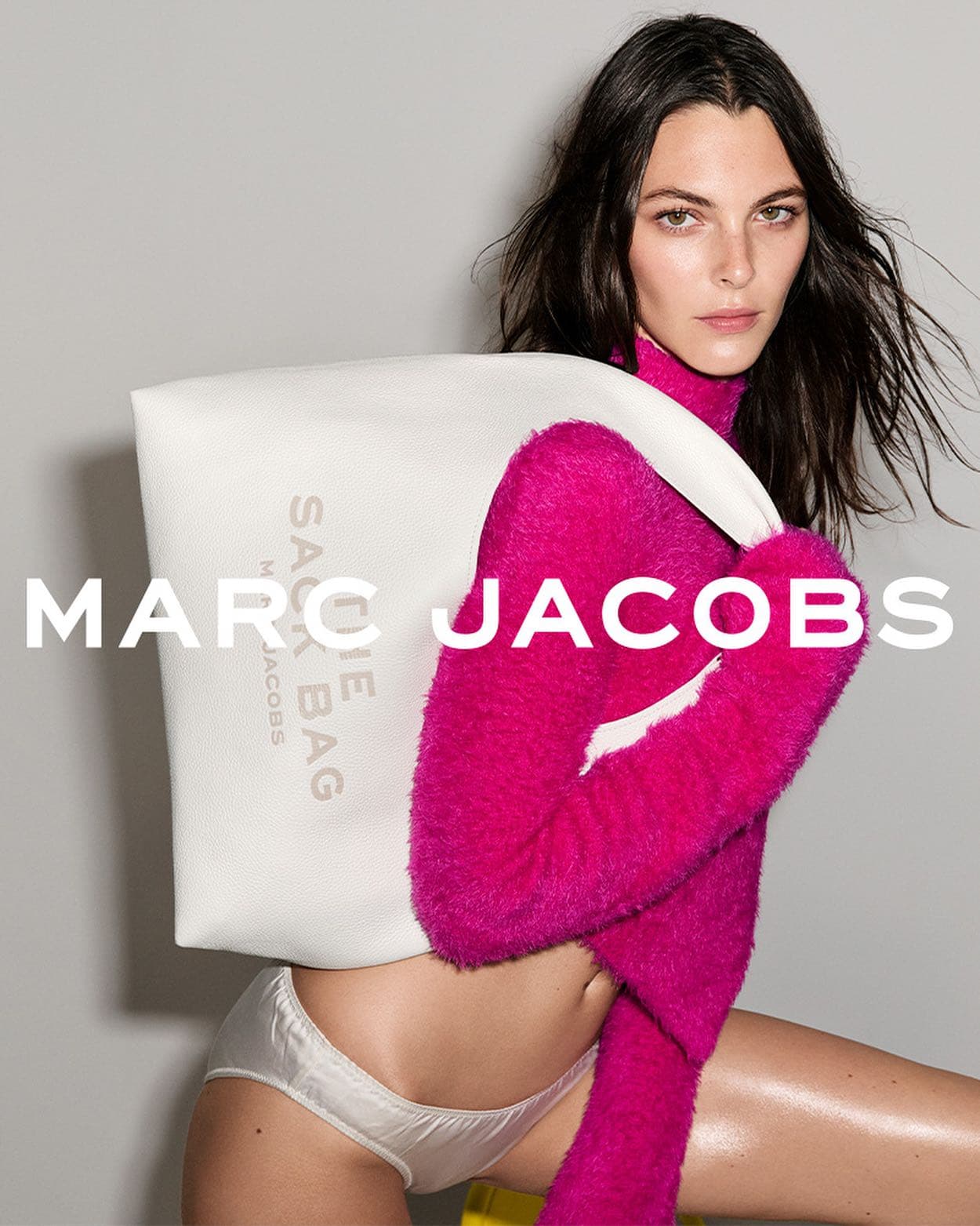 A fashionable adult female poses with a white tote bag with the logo 'Marc Jacobs The Sac Bag' in a studio shot with a gray background.