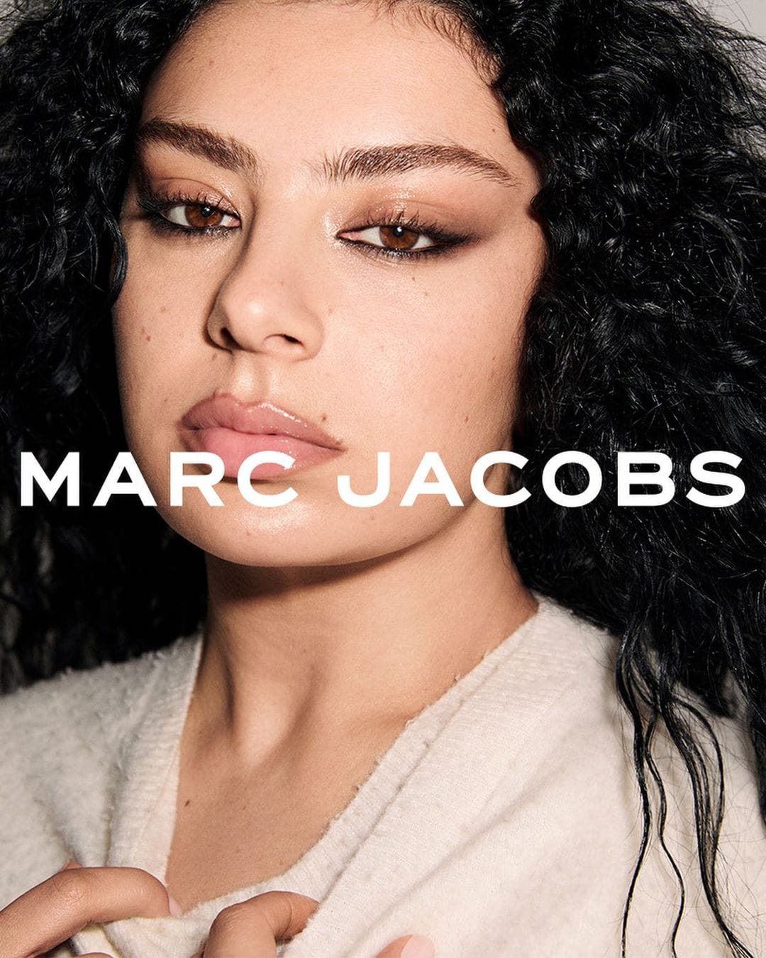 Close-up beauty shot of a young adult female model with curly hair wearing eye makeup and a cream sweater, featuring Marc Jacobs branding.