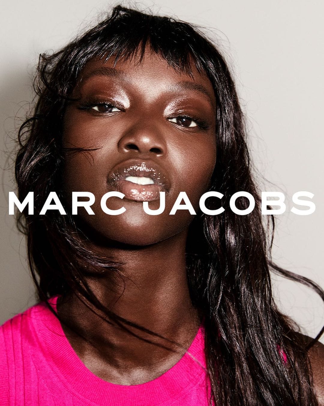 Close-up portrait of a dark-skinned woman with glossy makeup and long dark hair, wearing a pink top against a neutral backdrop.