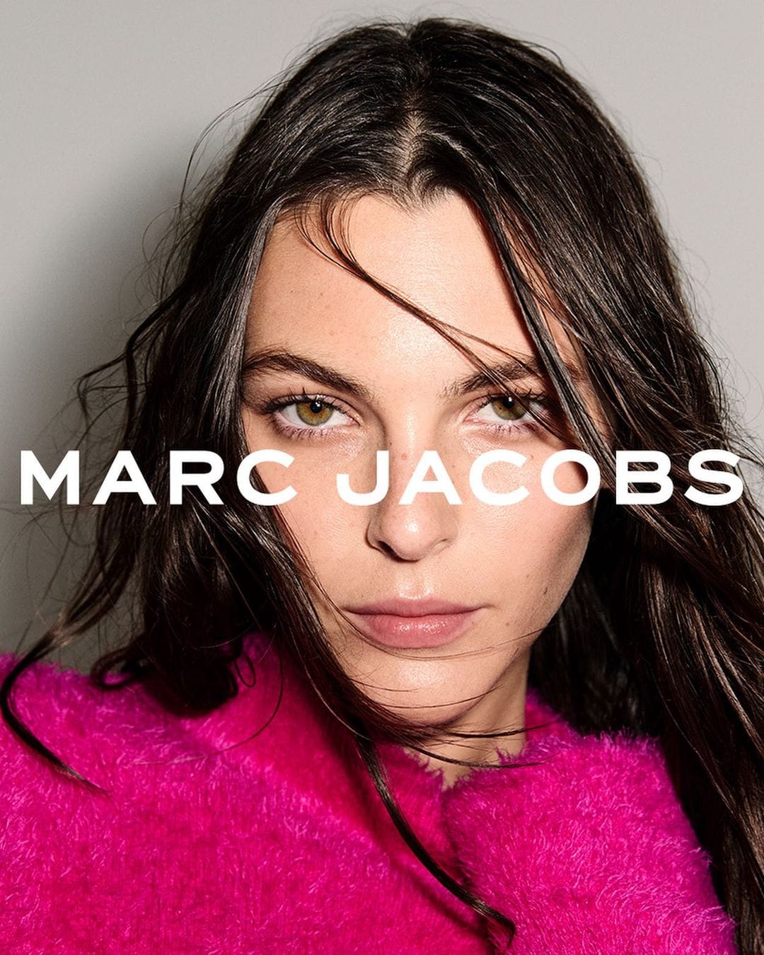 Close-up portrait of a brunette woman wearing a vibrant pink, fuzzy top with "MARC JACOBS" text overlay on a light gray background.