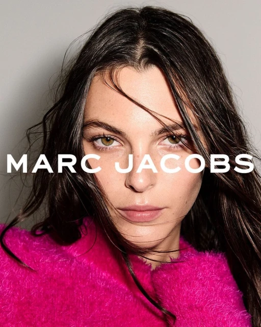Close-up portrait of a brunette woman wearing a vibrant pink, fuzzy top with "MARC JACOBS" text overlay on a light gray background.