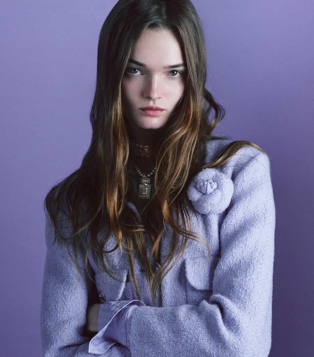 Portrait of a young woman with long brown hair wearing a lavender jacket with a floral embellishment against a lavender backdrop.