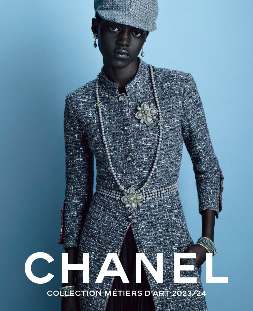 Fashion shot: a dark-skinned woman models a tweed overcoat and matching hat against a blue backdrop, showcasing an elegant, classic style.
