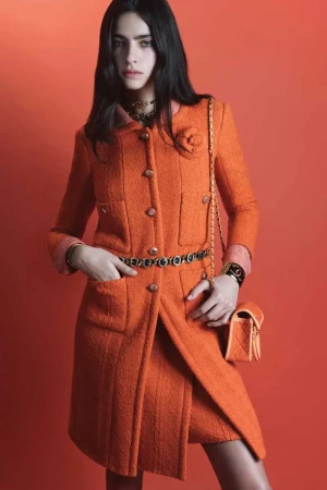 Fashionable woman in orange tweed coat and skirt, adorned with rose and chain-strap purse, poses against vibrant orange backdrop.