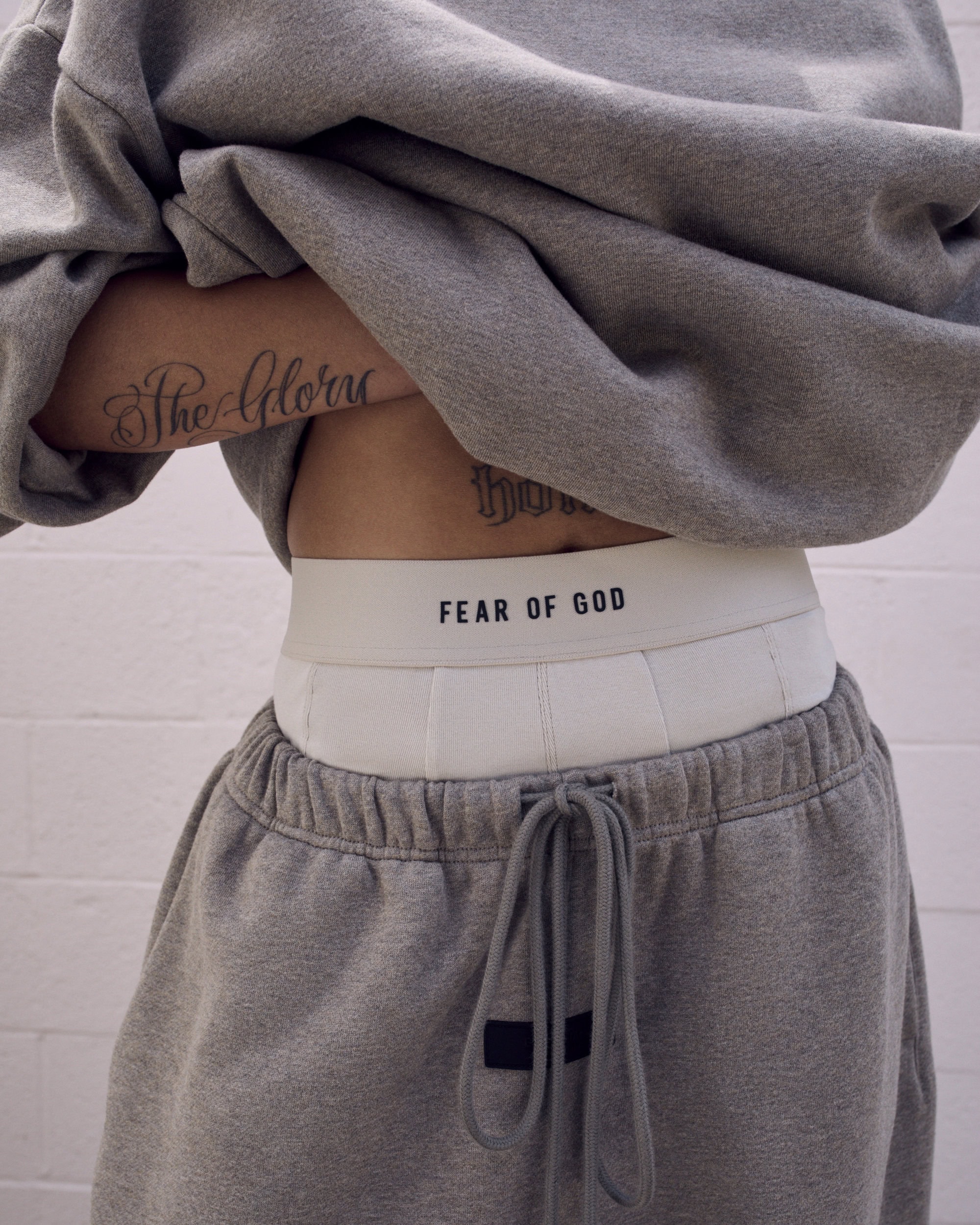 Waist up view of a person wearing a gray sweatshirt and sweatpants, white underwear band with 'FEAR OF GOD' text visible.