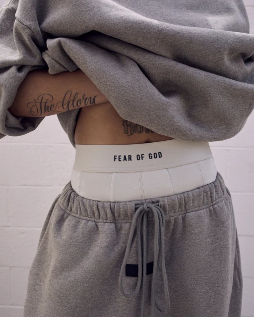 Waist up view of a person wearing a gray sweatshirt and sweatpants, white underwear band with 'FEAR OF GOD' text visible.