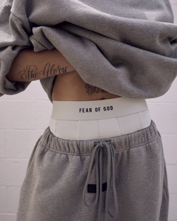 Waist up view of a person wearing a gray sweatshirt and sweatpants, white underwear band with 'FEAR OF GOD' text visible.