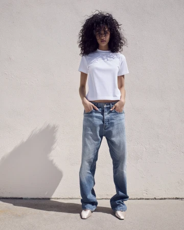 Model with long dark curly hair wearing a white t-shirt and blue jeans standing against a plain light wall
