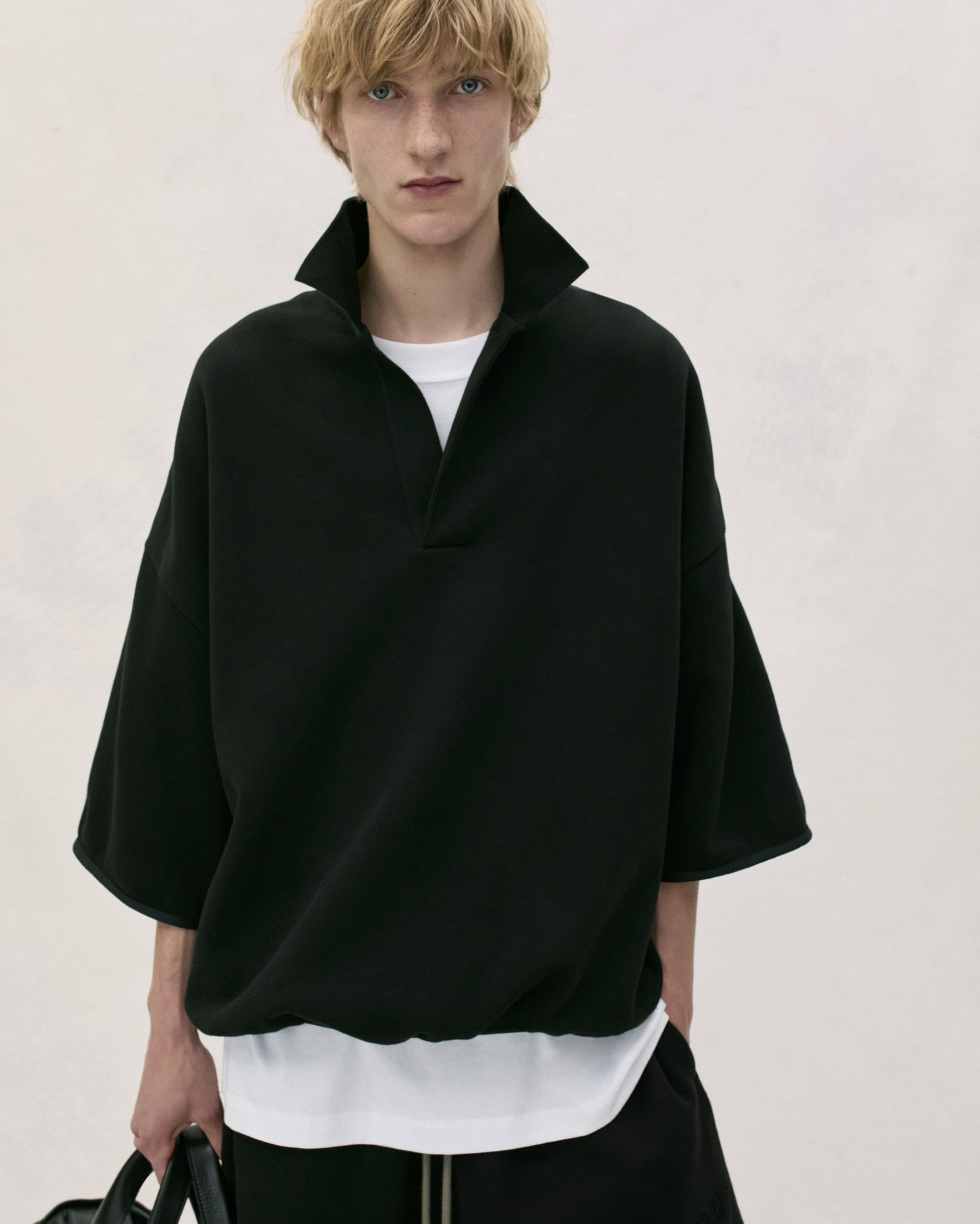 Model in black polo collar shirt and white t shirt standing against a neutral background for Fear of God.