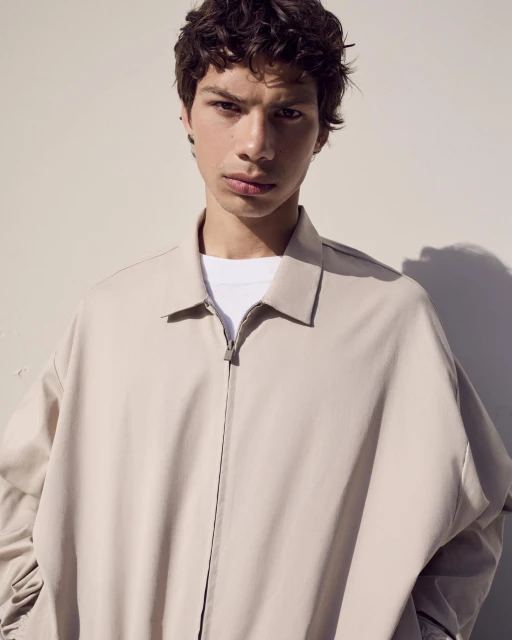 Portrait of a young adult male wearing a beige jacket and a white t-shirt standing against a light-colored wall, medium shot.