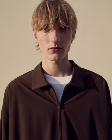 A fair-skinned young adult male with blond hair poses indoors wearing a brown zipper jacket, captured in a medium shot against a neutral background.