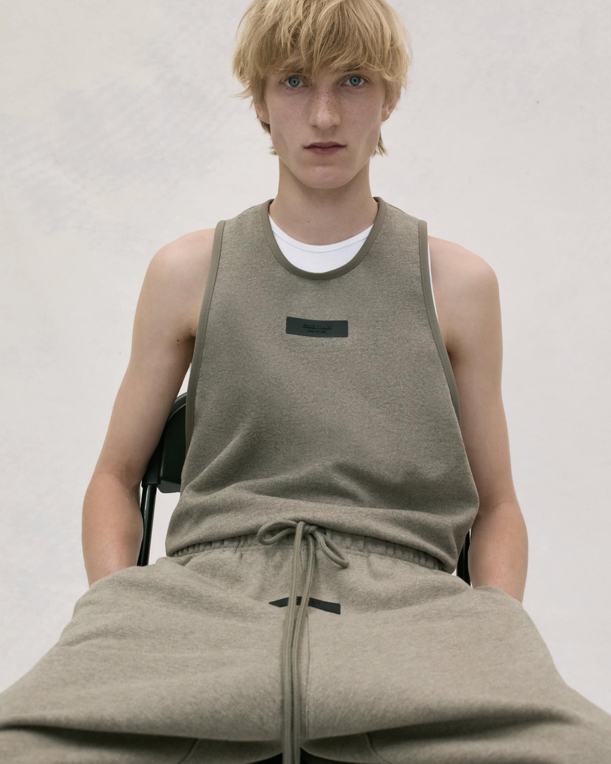 A fair-skinned young adult male sits on a chair, wearing an olive-green tank top and sweatpants, posing in a photo studio with bright lighting.