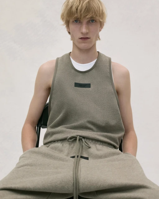 A fair-skinned young adult male sits on a chair, wearing an olive-green tank top and sweatpants, posing in a photo studio with bright lighting.