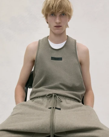 A fair-skinned young adult male sits on a chair, wearing an olive-green tank top and sweatpants, posing in a photo studio with bright lighting.