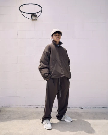 Young man wearing a brown jacket and matching track pants standing against a pale brick wall beneath a basketball hoop.
