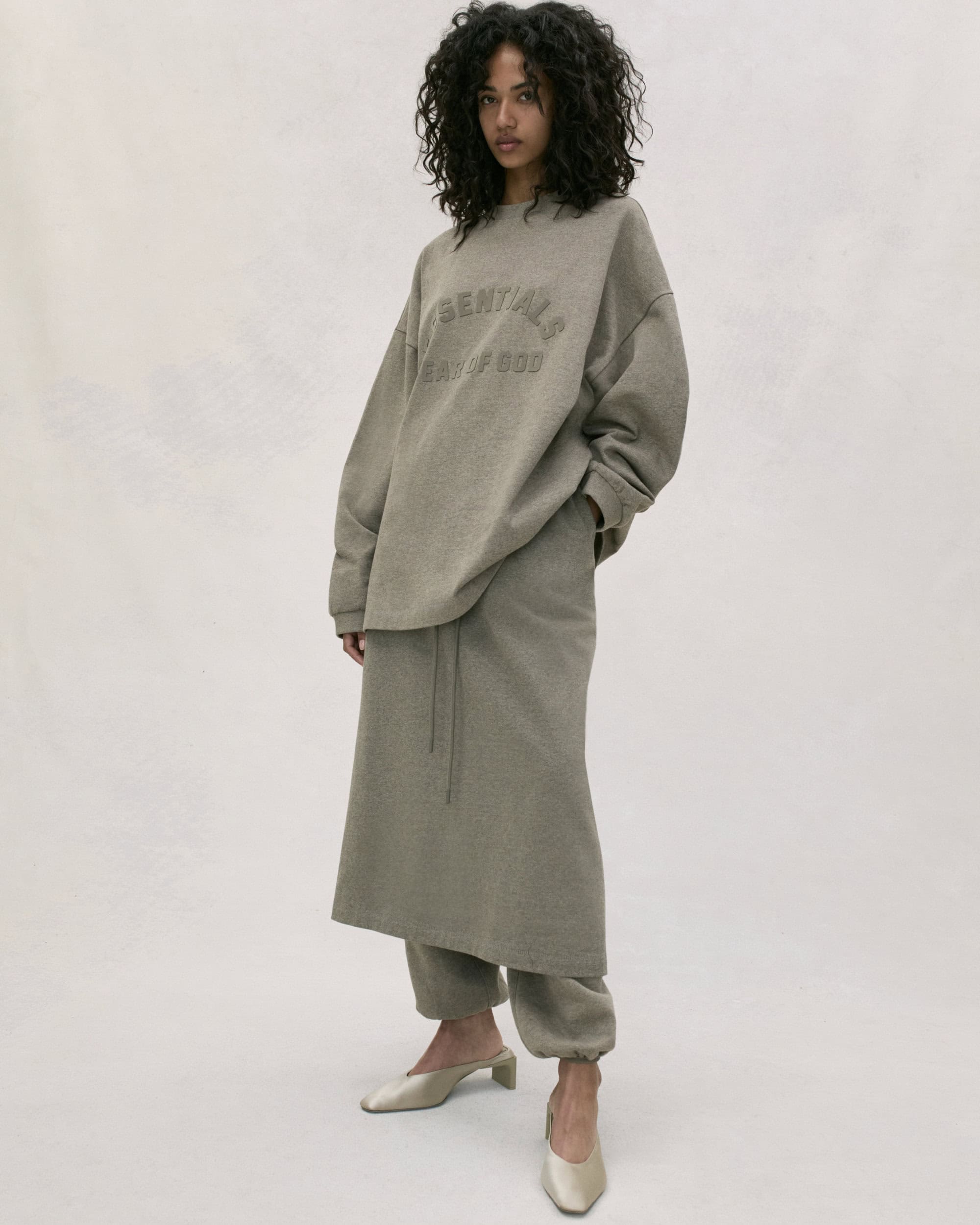 Adult woman with curly hair posing indoors in a gray sweater with text and a long skirt and mule shoes, against a neutral background.