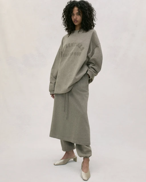 Adult woman with curly hair posing indoors in a gray sweater with text and a long skirt and mule shoes, against a neutral background.