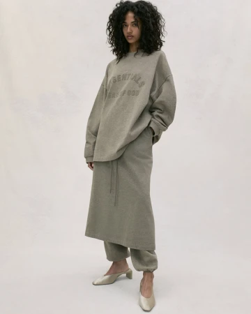 Adult woman with curly hair posing indoors in a gray sweater with text and a long skirt and mule shoes, against a neutral background.