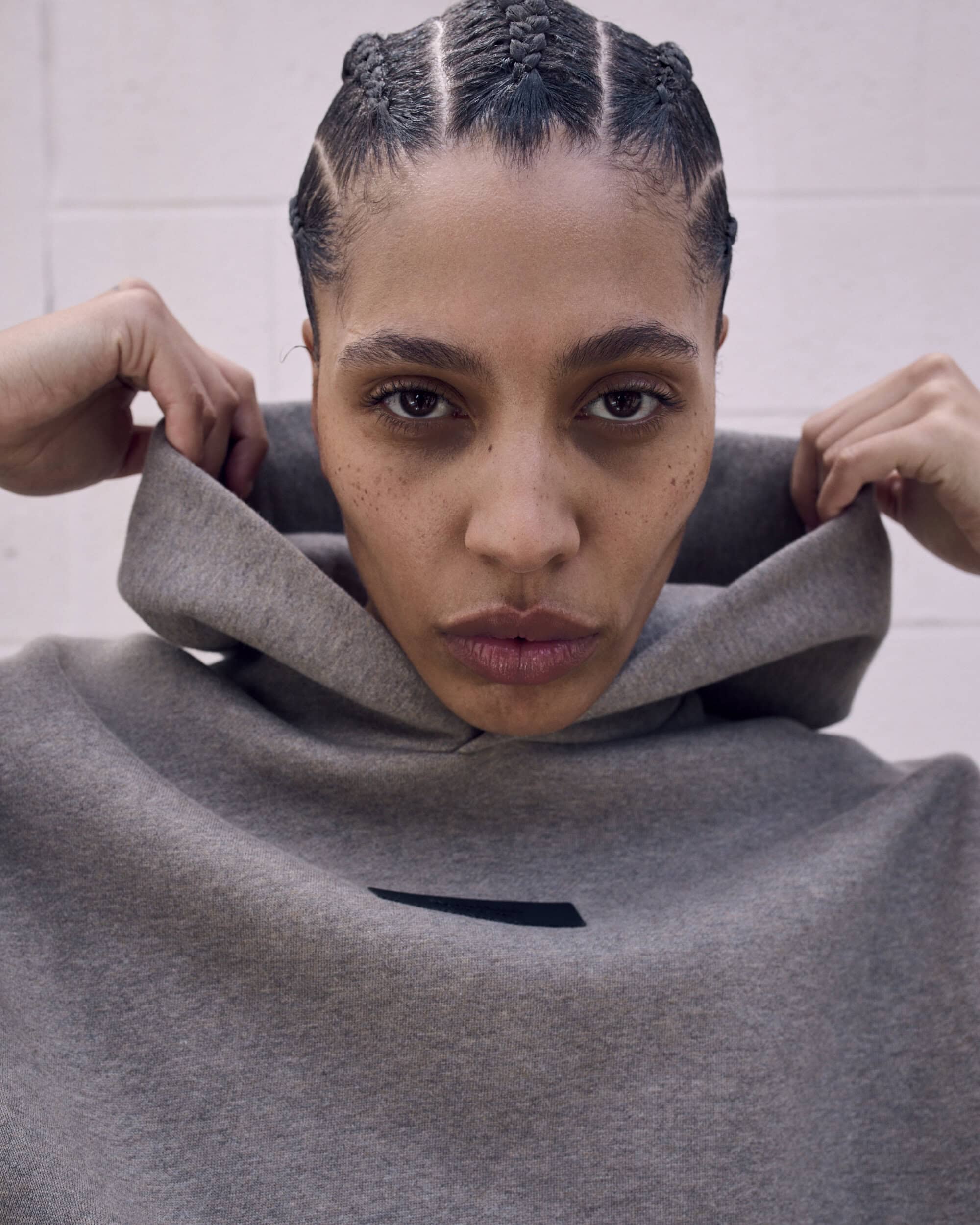 Close-up portrait of an adult female wearing a grey hoodie and a modern braided hairstyle against a neutral indoor background.