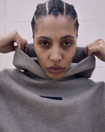 Close-up portrait of an adult female wearing a grey hoodie and a modern braided hairstyle against a neutral indoor background.