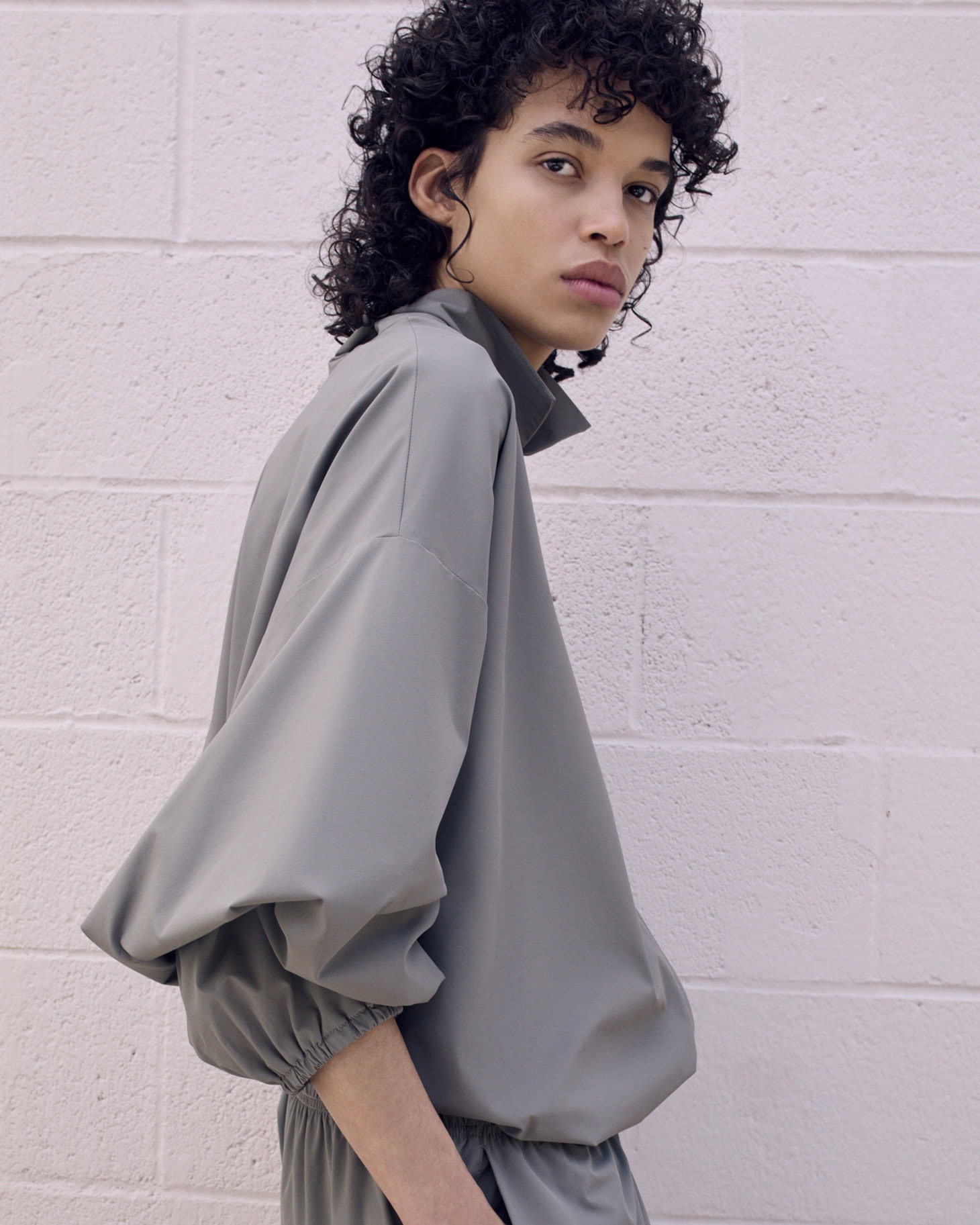 Fashionable adult with curly hair poses in a studio wearing a contemporary grey-green matching top and pants for a fashion shoot.