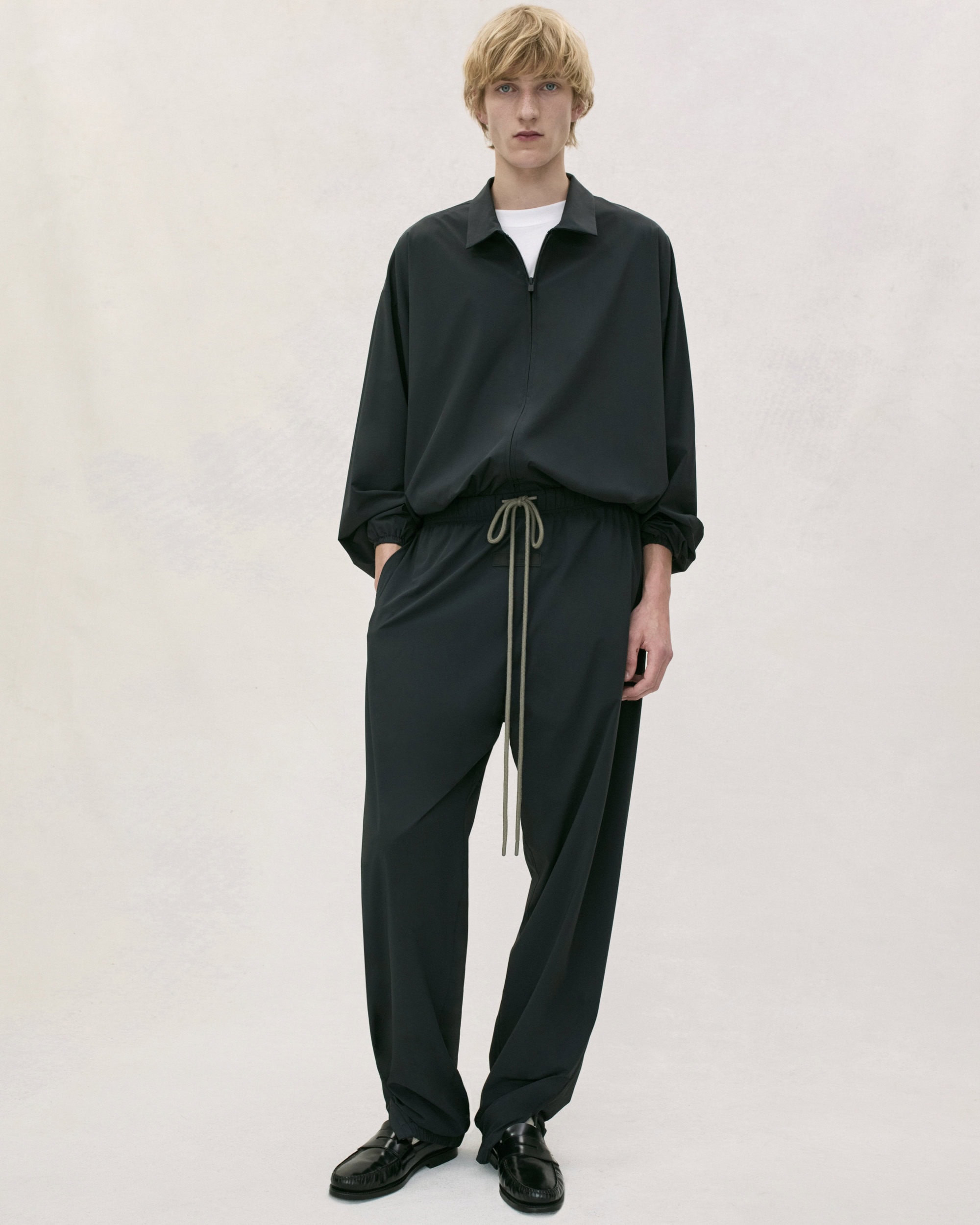 A male model standing against a light background wearing a dark zip-up jacket and relaxed trousers for Fear of God.