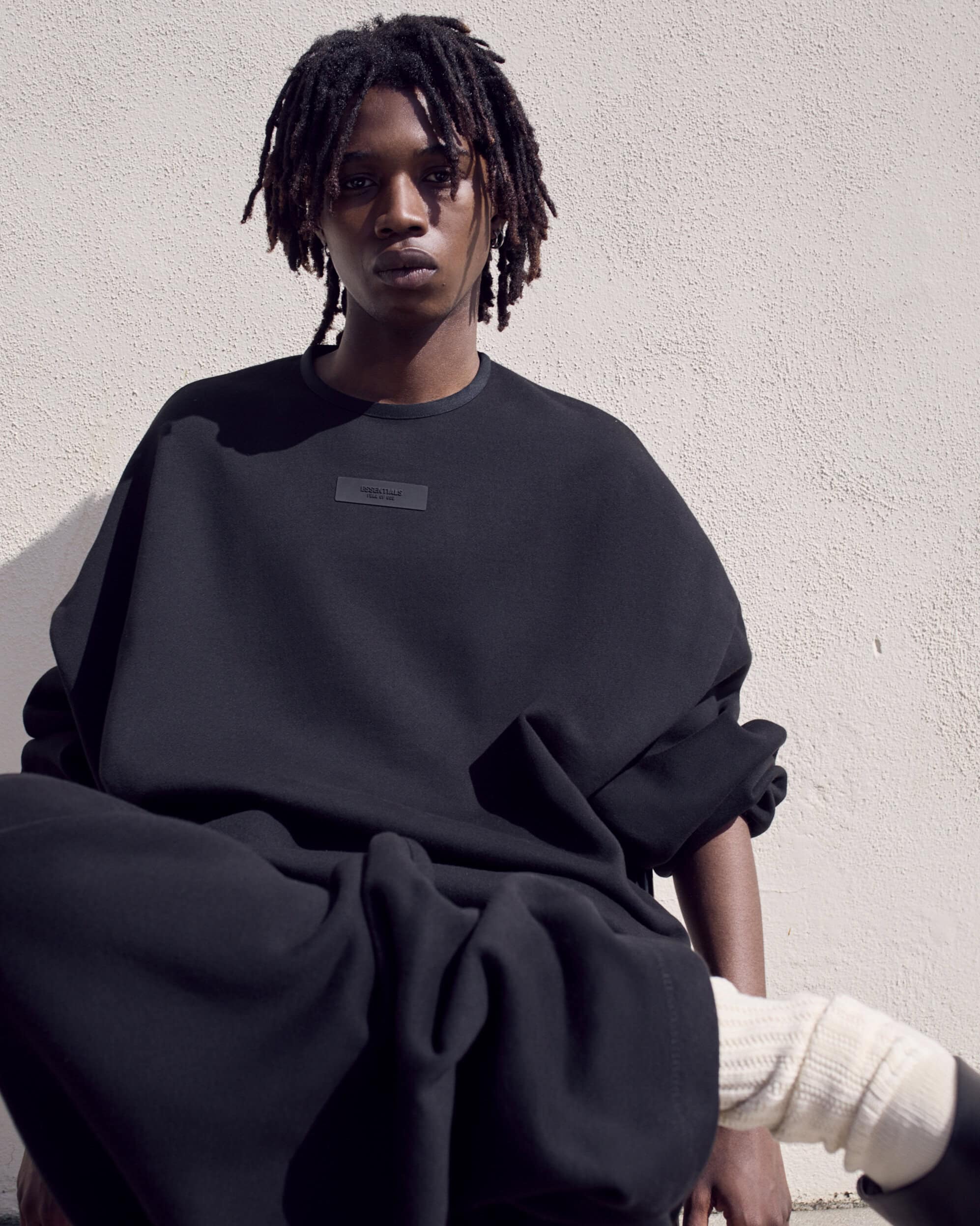 Studio shot of a young adult male with dreadlocks posing in a black, oversized sweatshirt and pants, set against a neutral background.