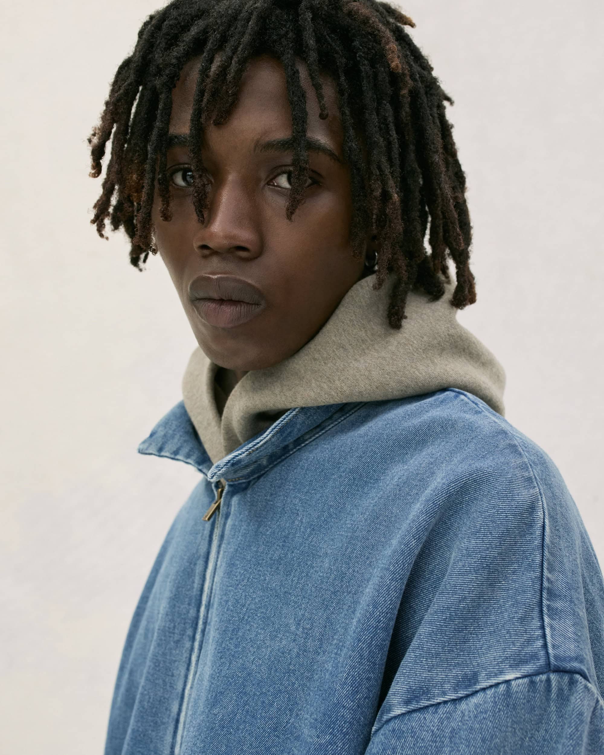 Close-up shot of a Black male model with dreadlocks wearing a blue denim jacket over a grey hoodie, posing in a photo studio.