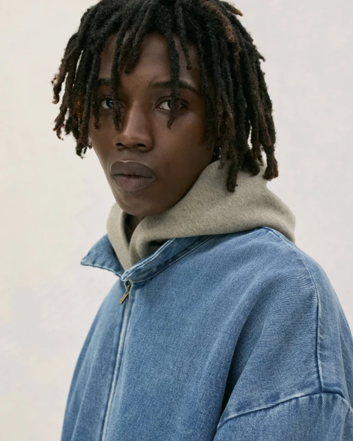 Close-up shot of a Black male model with dreadlocks wearing a blue denim jacket over a grey hoodie, posing in a photo studio.