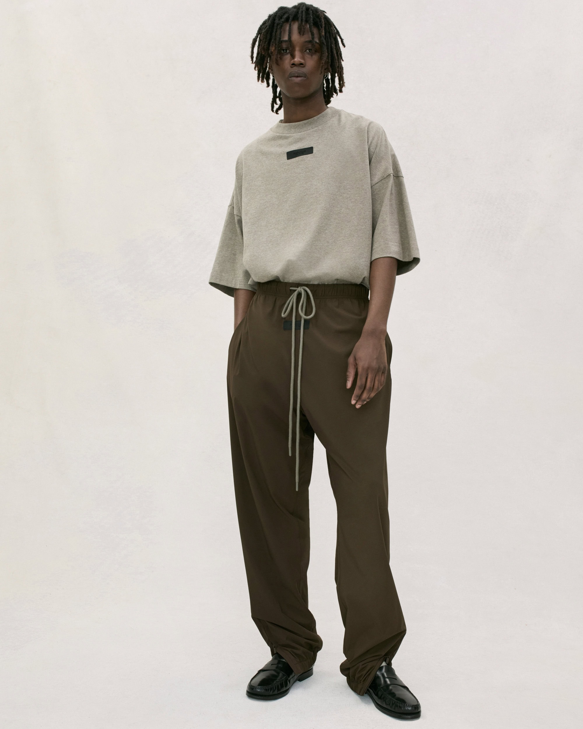 Adult male standing indoors wearing a beige t-shirt and brown drawstring pants in a studio with soft lighting.