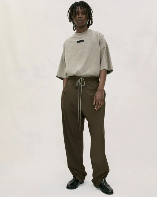 Adult male standing indoors wearing a beige t-shirt and brown drawstring pants in a studio with soft lighting.