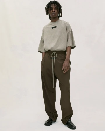 Adult male standing indoors wearing a beige t-shirt and brown drawstring pants in a studio with soft lighting.