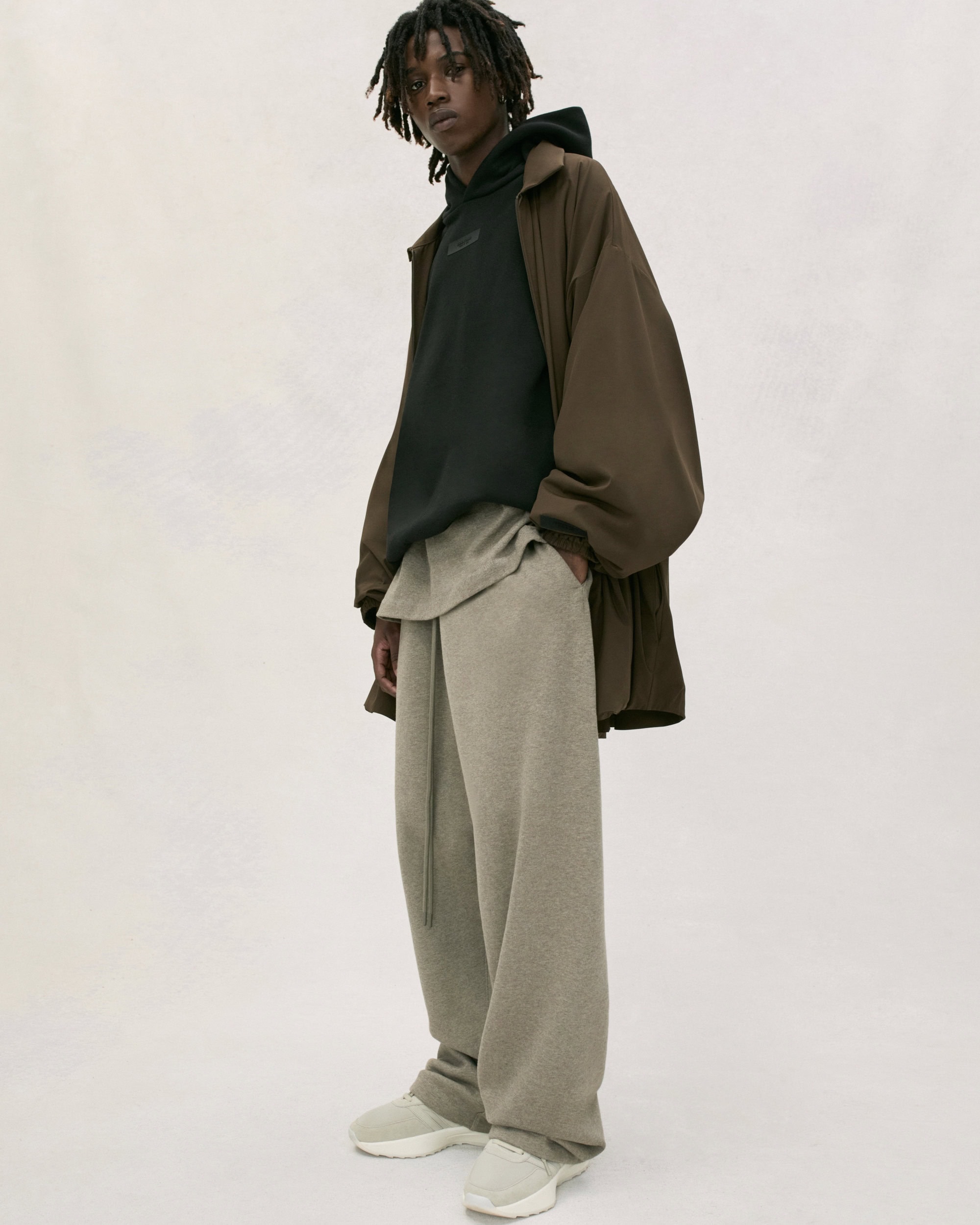 A person models brown wide-leg pants, sweatshirt, hoodie and jacket in a studio. The lighting is soft and neutral with a white background.