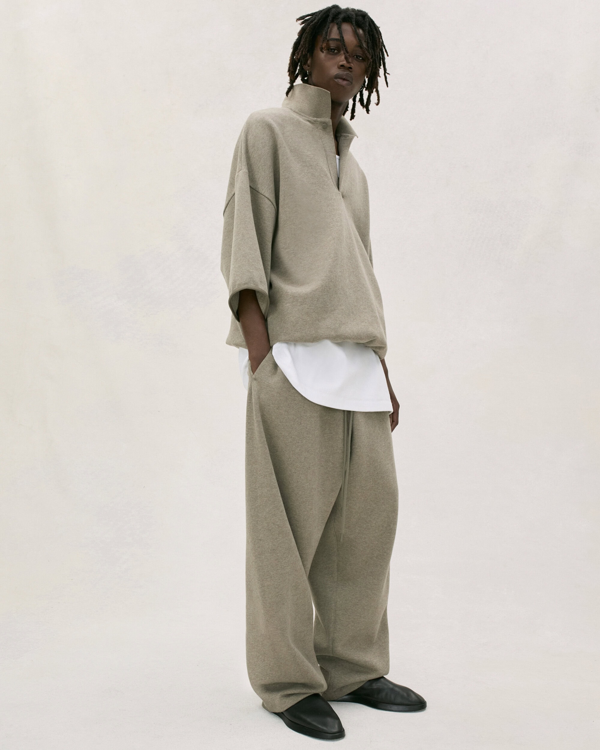 Fashion portrait of adult male model wearing beige quarter-zip sweatshirt and sweatpants with black shoes in a neutral studio setting.