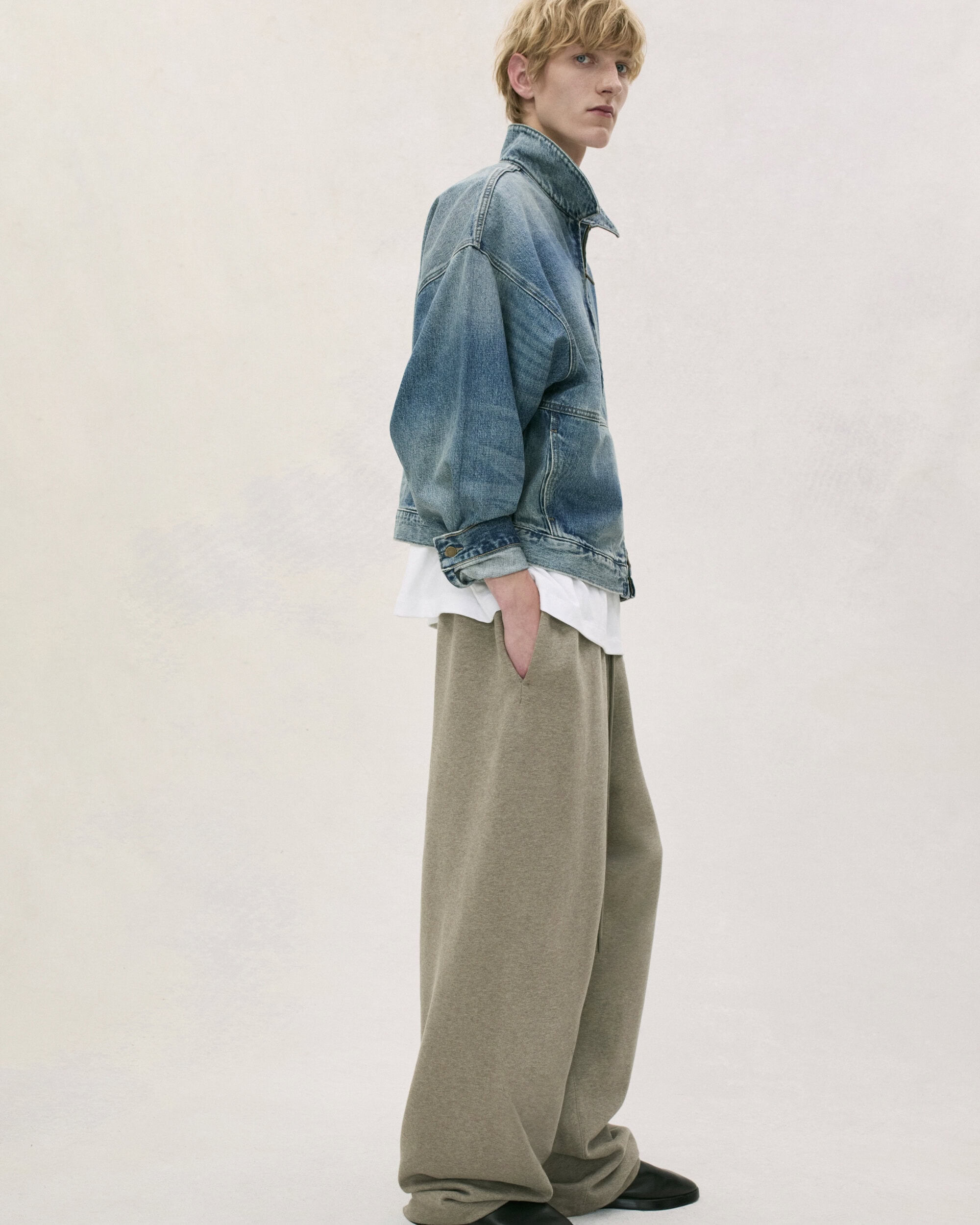 Adult male poses in a photo studio wearing a denim jacket, white t-shirt, and large grey sweatpants against an off-white background.