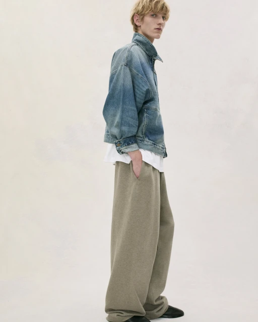 Adult male poses in a photo studio wearing a denim jacket, white t-shirt, and large grey sweatpants against an off-white background.