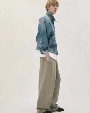 Adult male poses in a photo studio wearing a denim jacket, white t-shirt, and large grey sweatpants against an off-white background.