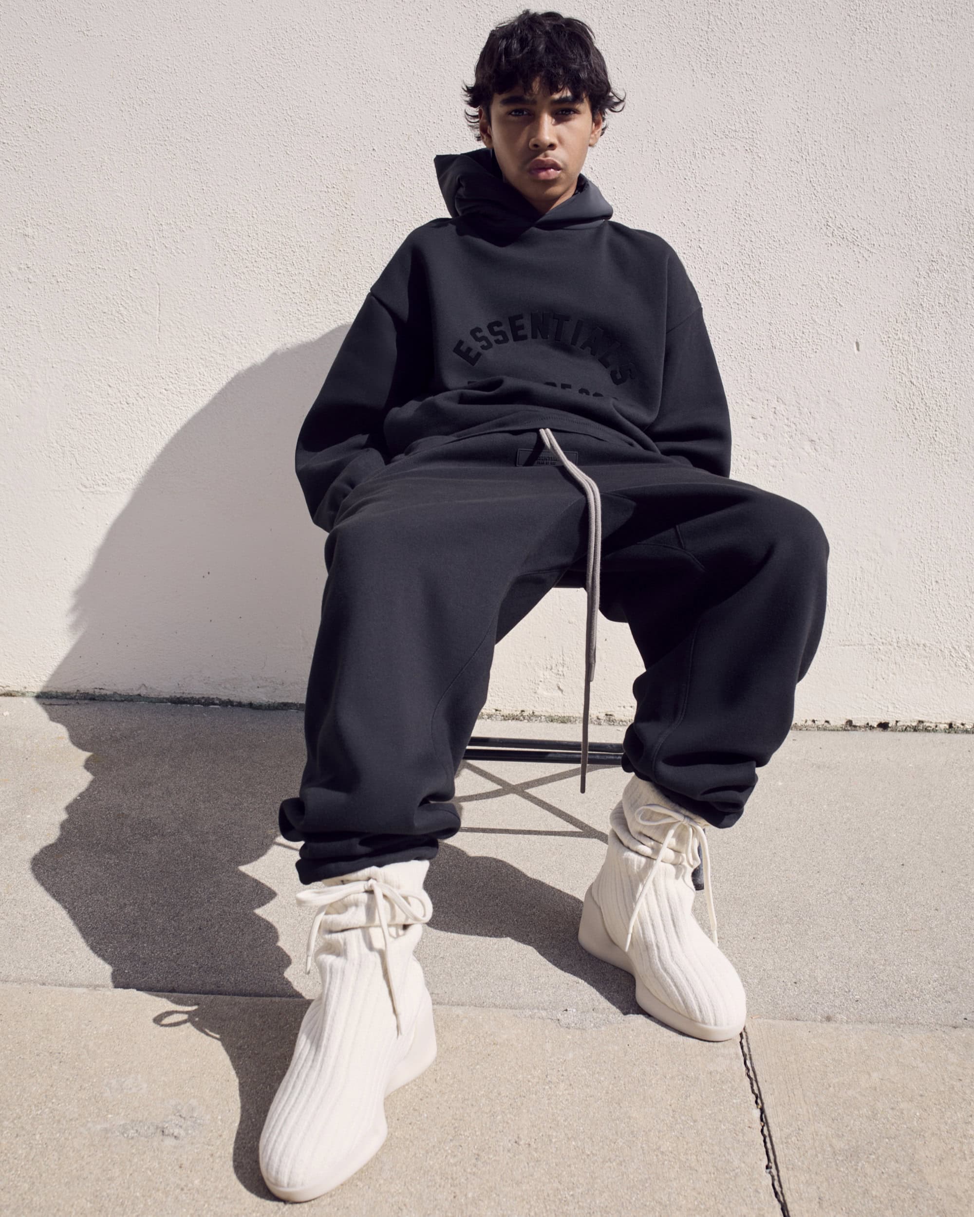 A person sits in black sweat suit with white knit shoes against a white wall in sunlight, casting shadows on the textured background.