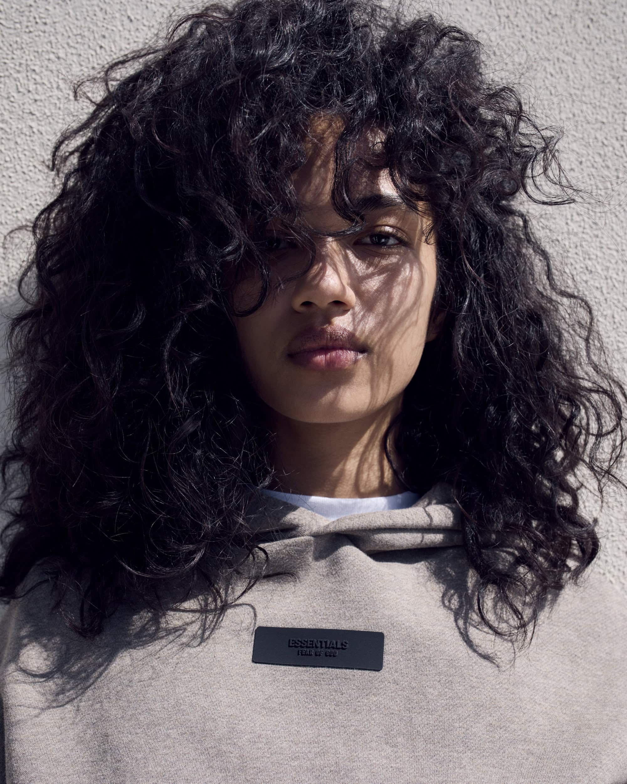 Close-up portrait of a young adult female with dark curly hair wearing a grey hoodie with an ESSENTIALS label, against a neutral wall.
