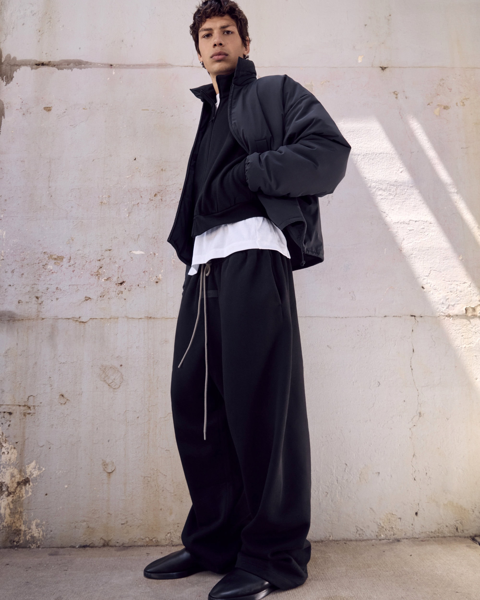 Male fashion model wears a black puffer jacket, white top, and black sweatpants in a bright studio with a concrete wall backdrop.